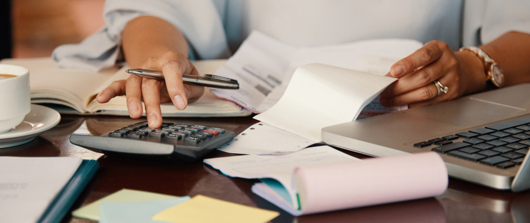 close em uma pessoa fazendo contas e, sobre a mesa, estão calculadora, computador, blocos de papéis e boletos
