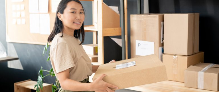 mulher sorri enquanto segura uma caixa de encomenda, ao lado estão várias outras caixas semelhantes