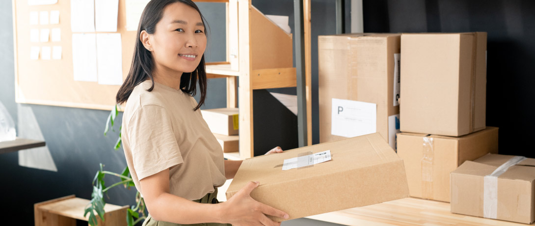 mulher sorri enquanto segura uma caixa de encomenda, ao lado estão várias outras caixas semelhantes