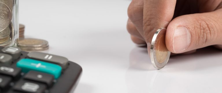 - Detalhe de uma mão segurando uma moeda ao lado de um pote de moedas e uma calcudadora