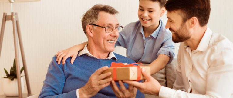 Homem mais velho sorrindo ao receber presente de Dia dos Pais de um homem mais jovem e uma criança.