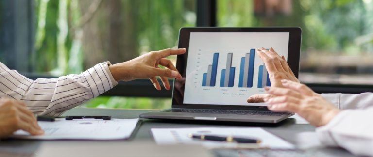 Duas pessoas analisando gráficos de barras em um notebook, representando a avaliação de indicadores de vendas.